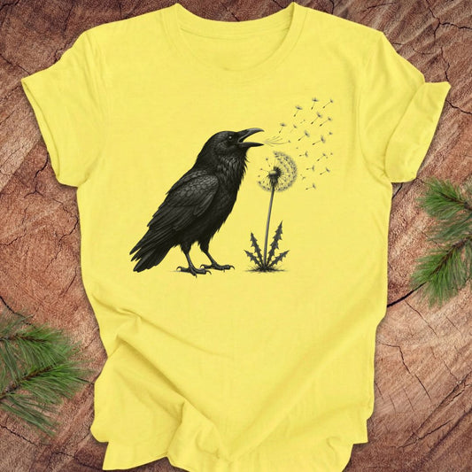 Yellow t-shirt with a raven and dandelion design on a wood background.