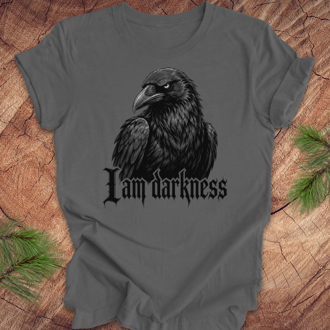 A dark gray t-shirt featuring a black raven graphic and the gothic text 'I am darkness' printed on the front, displayed flat on a rustic wood background with pine needles.