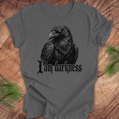 A dark gray t-shirt featuring a black raven graphic and the gothic text 'I am darkness' printed on the front, displayed flat on a rustic wood background with pine needles.