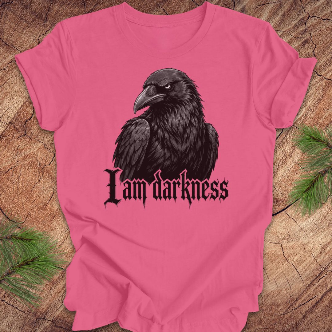 A pink short-sleeve t-shirt with a graphic of a black raven and the gothic text 'I am darkness' printed on the front, displayed flat on a rustic wood background with pine needles.