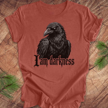 A terracotta-colored short-sleeve t-shirt featuring a black raven graphic and the gothic text 'I am darkness' printed on the front, laid flat on a rustic wood background with pine needles in the corners.
