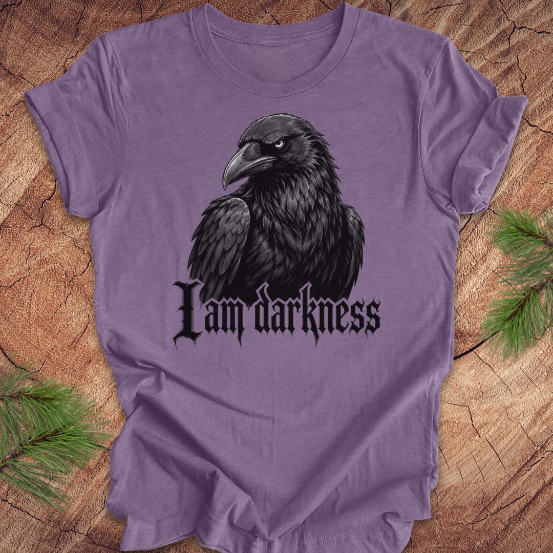 A purple short-sleeve t-shirt featuring a black raven graphic and the gothic text 'I am darkness' on the front, laid flat on a rustic wood background with pine needles in the corners.