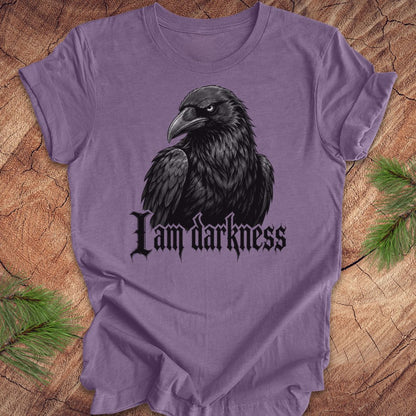 A purple short-sleeve t-shirt featuring a black raven graphic and the gothic text 'I am darkness' on the front, laid flat on a rustic wood background with pine needles in the corners.