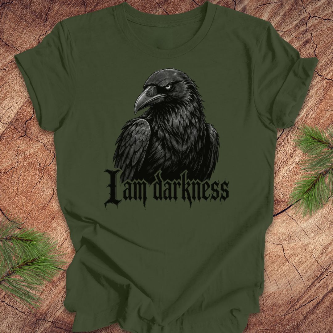An olive green short-sleeve t-shirt featuring a graphic of a black raven with the gothic text 'I am darkness' underneath, displayed flat on a rustic wood background with pine needles.
