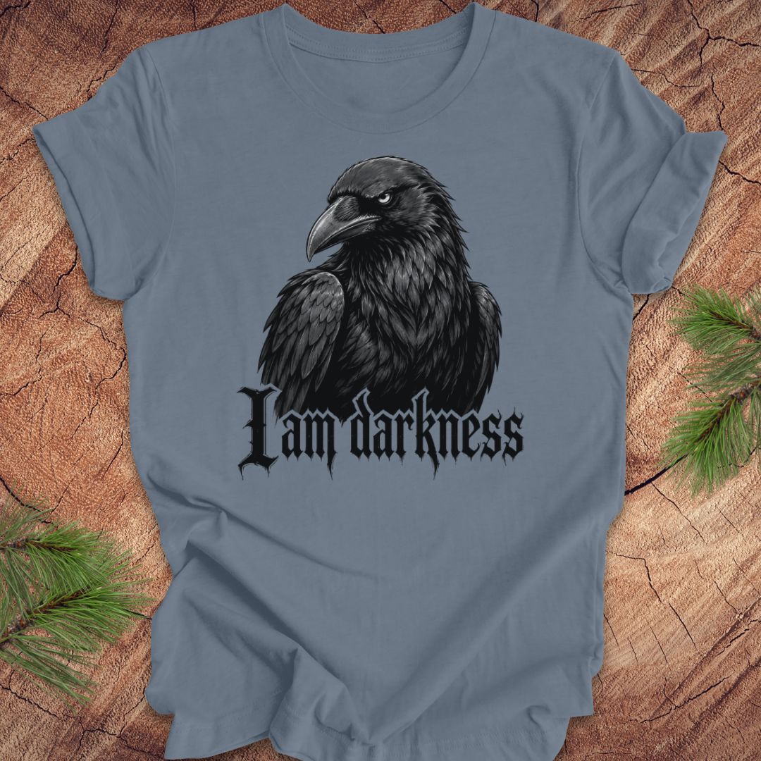 A slate blue t-shirt featuring a detailed black raven graphic and the gothic text 'I am darkness' printed on the front, displayed flat on a rustic wood background with pine needles.