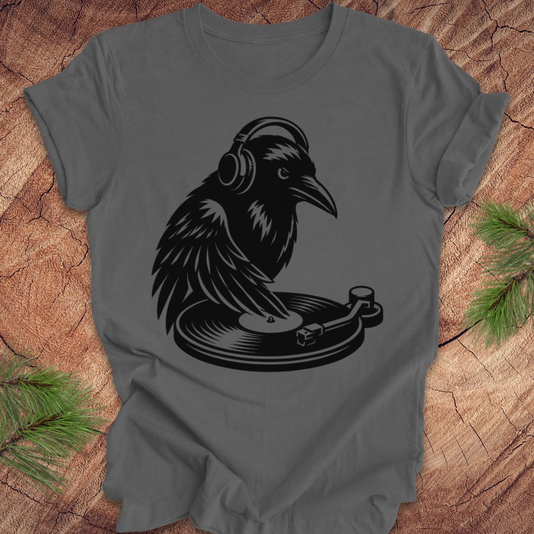 A dark gray crewneck t-shirt featuring a black graphic of a raven wearing headphones and playing a vinyl record, laid flat on a rustic wood background with pine needles in the corners.