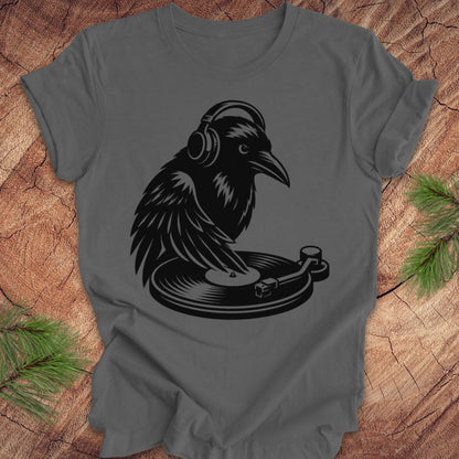 A dark gray crewneck t-shirt featuring a black graphic of a raven wearing headphones and playing a vinyl record, laid flat on a rustic wood background with pine needles in the corners.