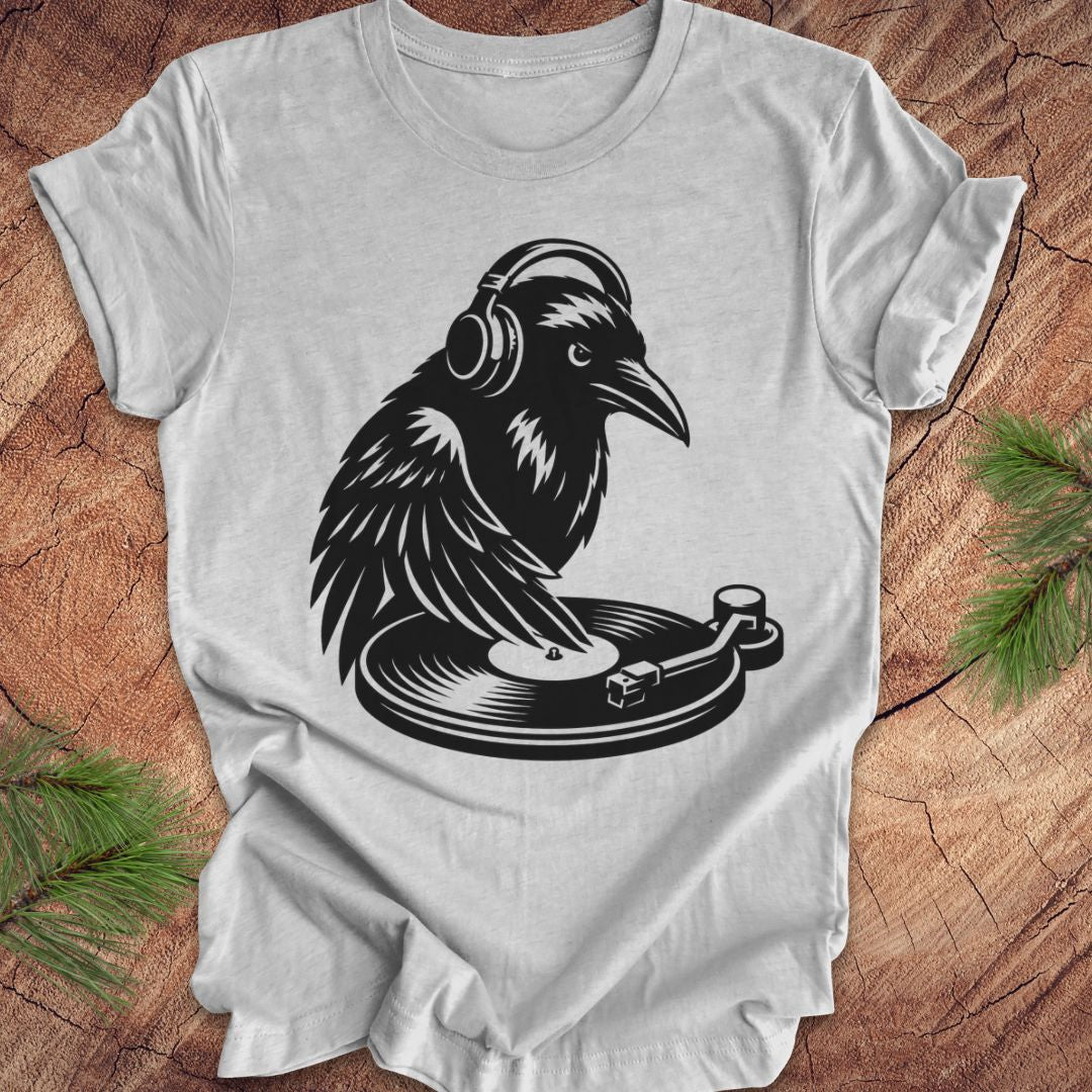 A light gray short-sleeve t-shirt laid flat on a rustic wood surface, featuring a black graphic of a raven wearing headphones and resting on a turntable. Green pine branches are placed in the corners as styling props.