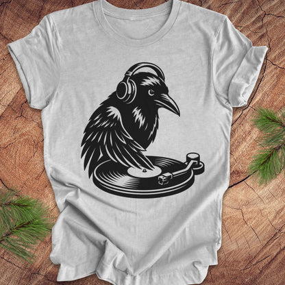 A light gray short-sleeve t-shirt laid flat on a rustic wood surface, featuring a black graphic of a raven wearing headphones and resting on a turntable. Green pine branches are placed in the corners as styling props.