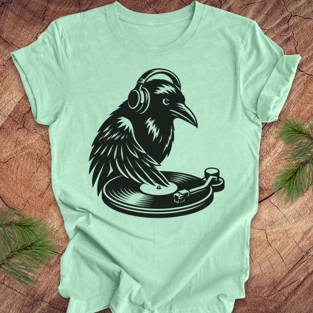 A mint green short-sleeve t-shirt laid flat on a rustic wood surface, featuring a black graphic of a raven wearing headphones and spinning a record player.