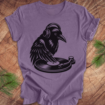 A purple short-sleeve t-shirt laid flat on a rustic wood surface, featuring a black graphic of a raven wearing headphones and sitting on a turntable. Green pine branches are placed in the corners of the frame.