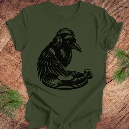 An olive green short-sleeve t-shirt with a black graphic of a raven wearing headphones and sitting on a turntable, laid flat on a rustic wood background with pine branches in the corners.