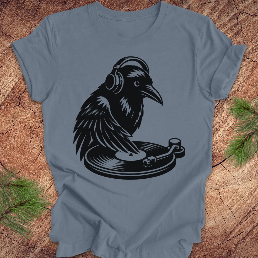 A slate blue crewneck t-shirt featuring a black graphic of a raven wearing headphones and spinning a record player. The t-shirt is laid flat on a rustic wood background with pine needles visible in the corners.
