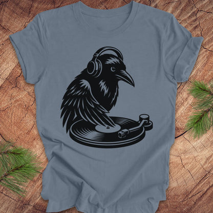 A slate blue crewneck t-shirt featuring a black graphic of a raven wearing headphones and spinning a record player. The t-shirt is laid flat on a rustic wood background with pine needles visible in the corners.