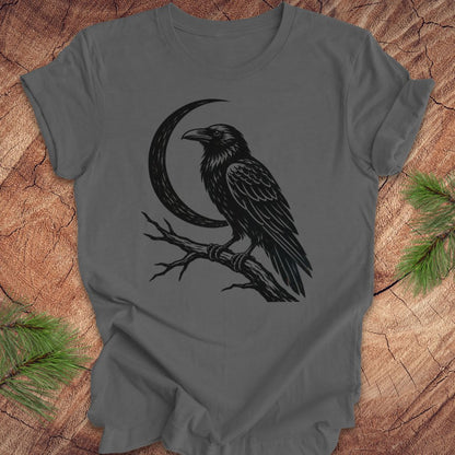 A dark gray crewneck t-shirt featuring a black graphic of a raven perched on a branch next to a crescent moon, laid flat on a rustic wood background with pine needles in the corners.
