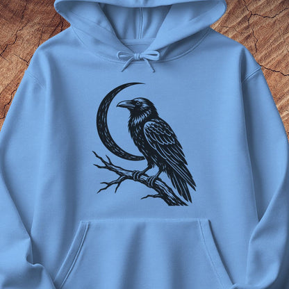 A light blue pullover hoodie laid flat on a rustic wood background, featuring a black graphic print of a raven perched on a branch in front of a crescent moon.