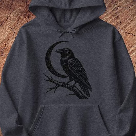 A dark heather gray hoodie featuring a black graphic print of a raven perched on a branch in front of a crescent moon, displayed against a rustic wood background.