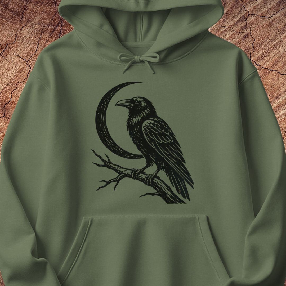 An olive green pullover hoodie featuring a black graphic print of a raven perched on a branch next to a crescent moon, displayed against a textured brown wood background.
