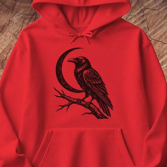 A bright red pullover hoodie with a black graphic of a raven perched on a branch in front of a crescent moon, displayed on a textured wood background.