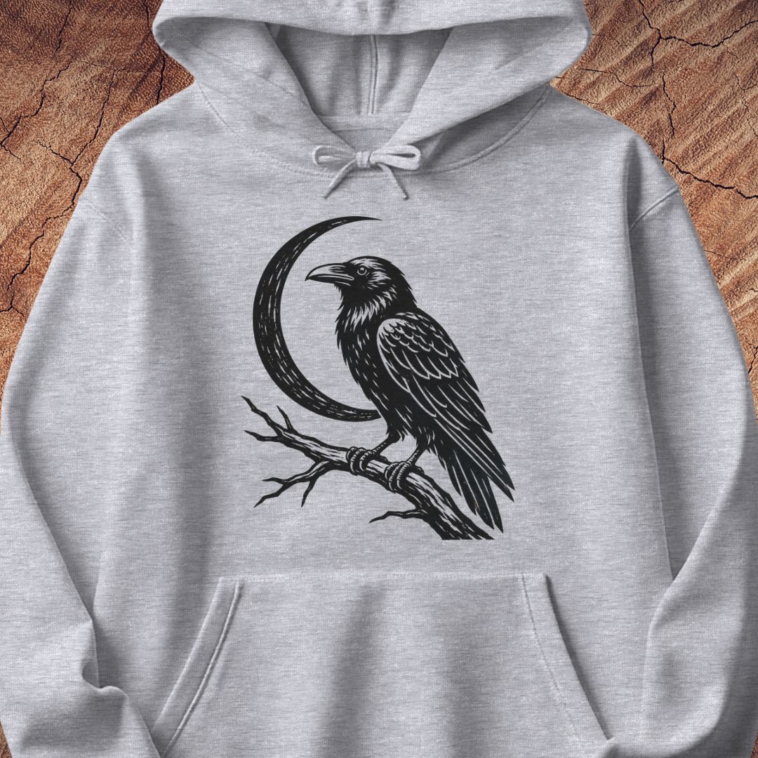 A light heather gray pullover hoodie featuring a black graphic print of a raven perched on a branch in front of a crescent moon, displayed flat against a rustic wood background.