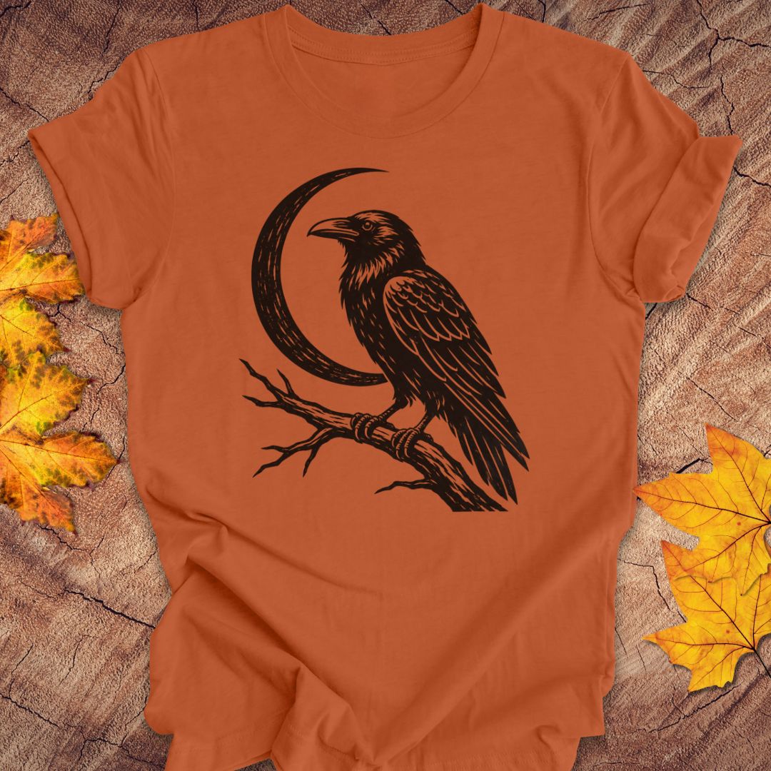 An orange short-sleeve t-shirt with a black graphic of a raven perched on a branch in front of a crescent moon, laid flat on a rustic wood background with autumn leaves.