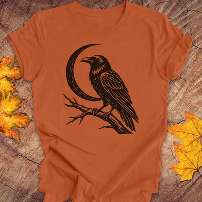 An orange short-sleeve t-shirt with a black graphic of a raven perched on a branch in front of a crescent moon, laid flat on a rustic wood background with autumn leaves.