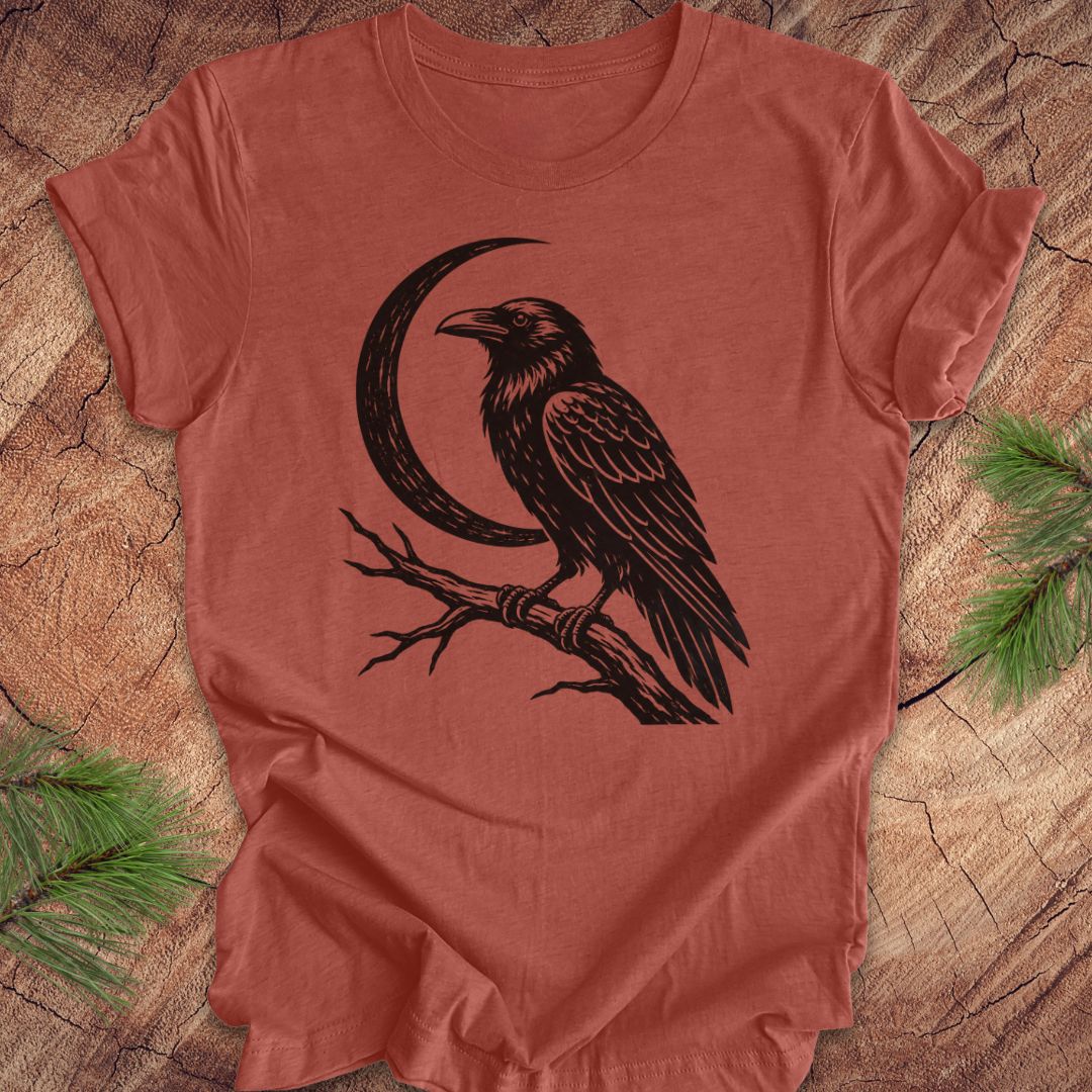 A terracotta-colored short-sleeve t-shirt with a black graphic of a raven perched on a branch next to a crescent moon, laid flat on a rustic wood background with pine needles in the corners.