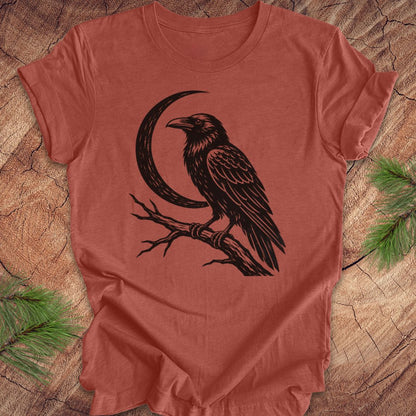 A terracotta-colored short-sleeve t-shirt with a black graphic of a raven perched on a branch next to a crescent moon, laid flat on a rustic wood background with pine needles in the corners.