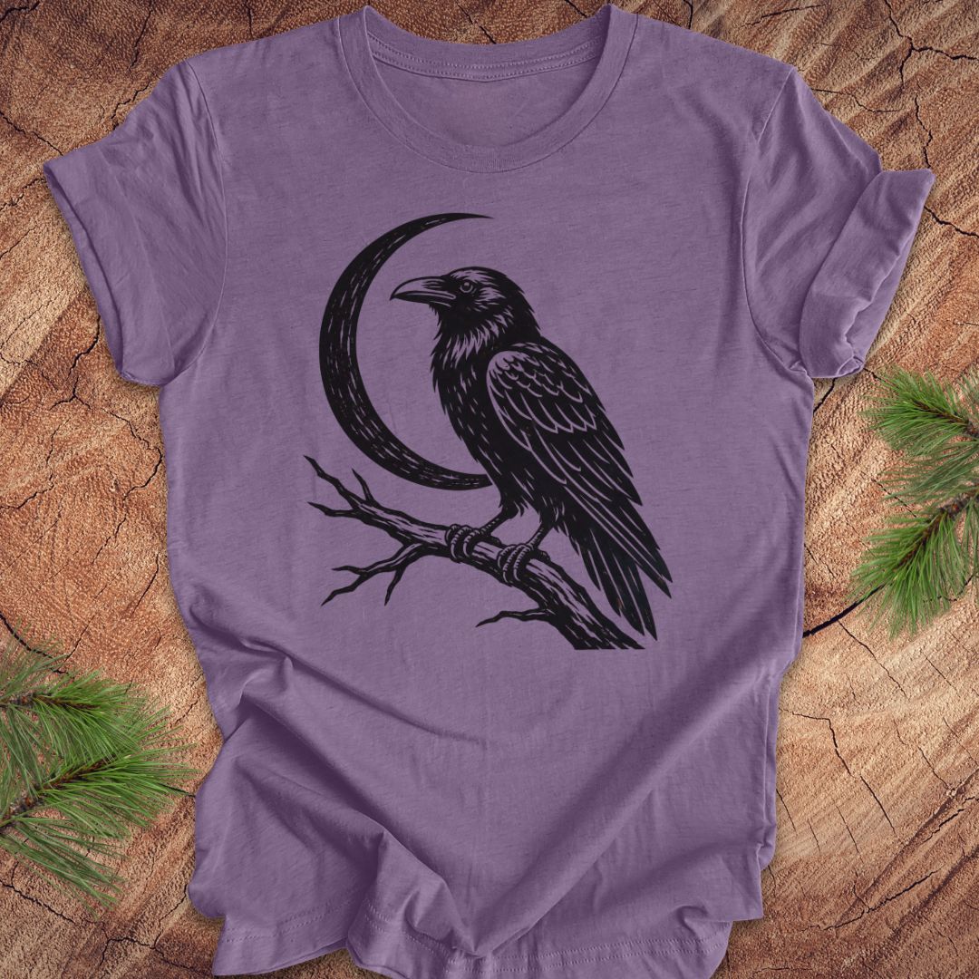 A lavender purple short-sleeve t-shirt with a black graphic of a raven perched on a branch next to a crescent moon, laid flat on a rustic wood background with pine needles in the corners.