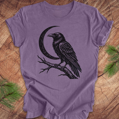 A lavender purple short-sleeve t-shirt with a black graphic of a raven perched on a branch next to a crescent moon, laid flat on a rustic wood background with pine needles in the corners.