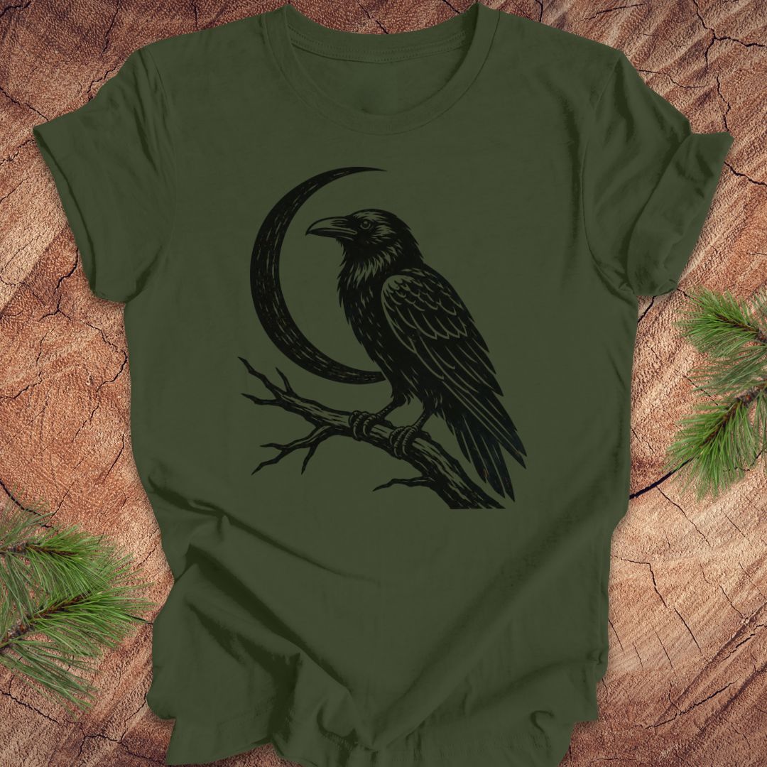 An olive green short-sleeve t-shirt with a black graphic of a raven perched on a branch in front of a crescent moon, laid flat on a rustic wood background with pine needles in the corners.
