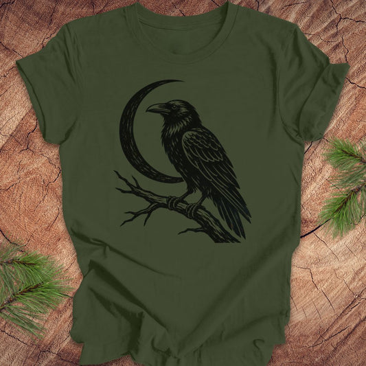An olive green short-sleeve t-shirt with a black graphic of a raven perched on a branch in front of a crescent moon, laid flat on a rustic wood background with pine needles in the corners.