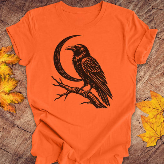 An orange short-sleeve crewneck t-shirt featuring a black graphic of a raven perched on a branch next to a crescent moon, laid flat on a rustic wood surface with yellow autumn leaves.