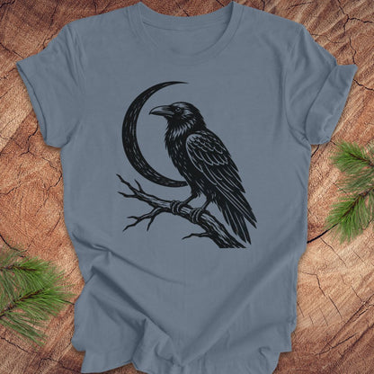 A slate blue crewneck t-shirt featuring a black graphic of a raven perched on a branch next to a crescent moon, laid flat on a rustic wood background with pine needles in the corners.