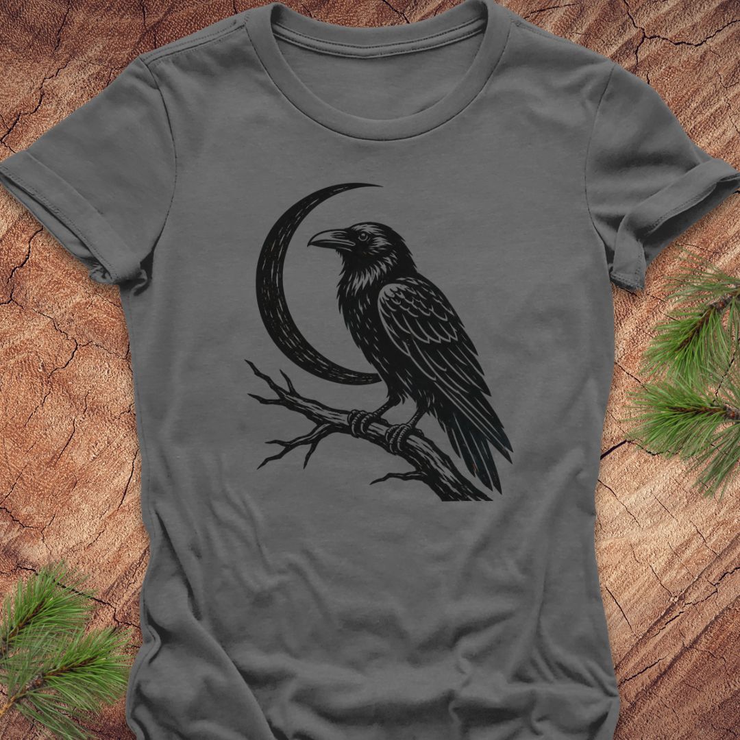 A dark gray short-sleeve t-shirt with a black graphic of a raven perched on a branch in front of a crescent moon, laid flat on a rustic wood background with pine needles in the corners.