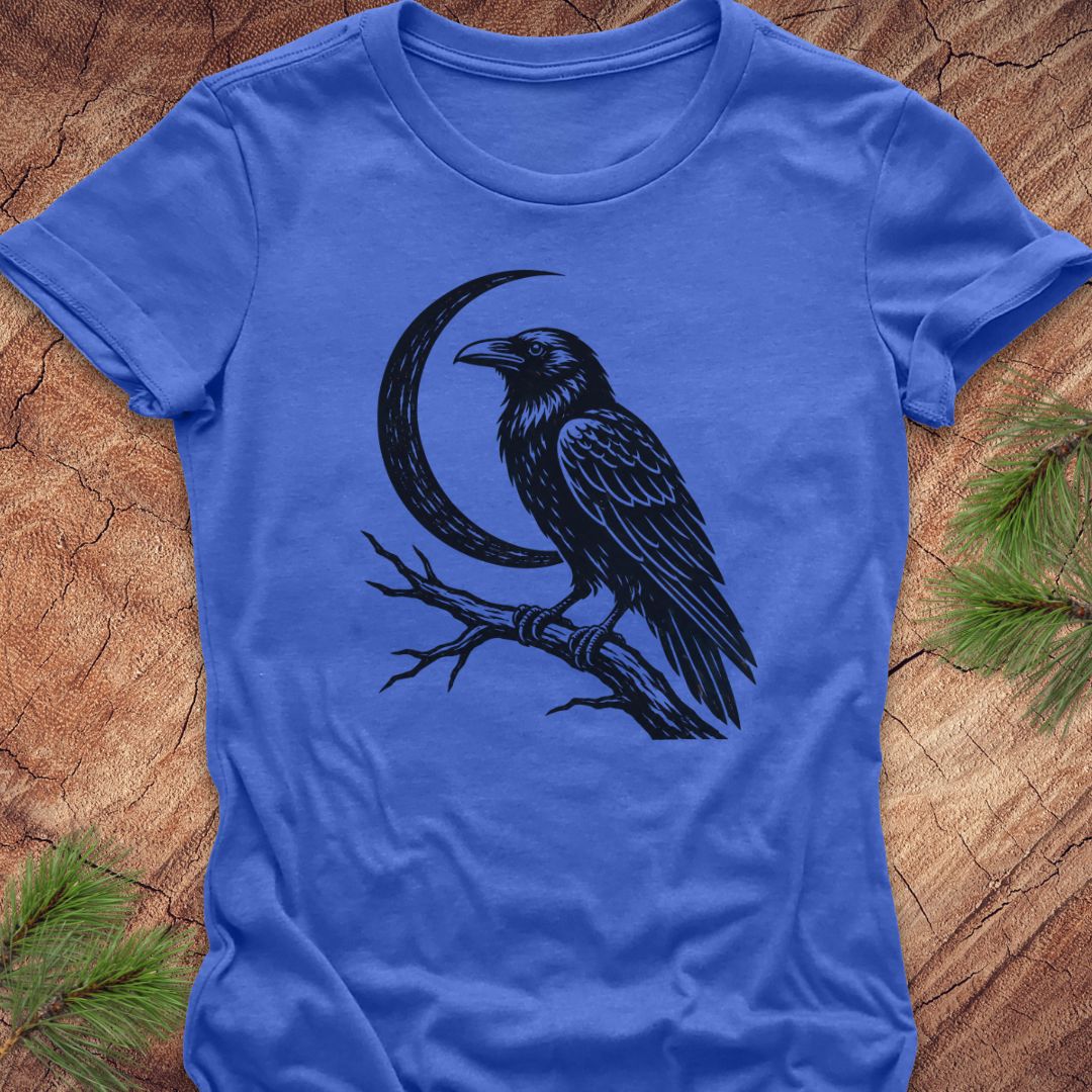 A royal blue short-sleeve t-shirt with a black graphic of a raven perched on a branch in front of a crescent moon, laid flat on a textured wood background with pine needles in the corners.