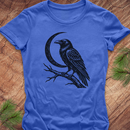 A royal blue short-sleeve t-shirt with a black graphic of a raven perched on a branch in front of a crescent moon, laid flat on a textured wood background with pine needles in the corners.