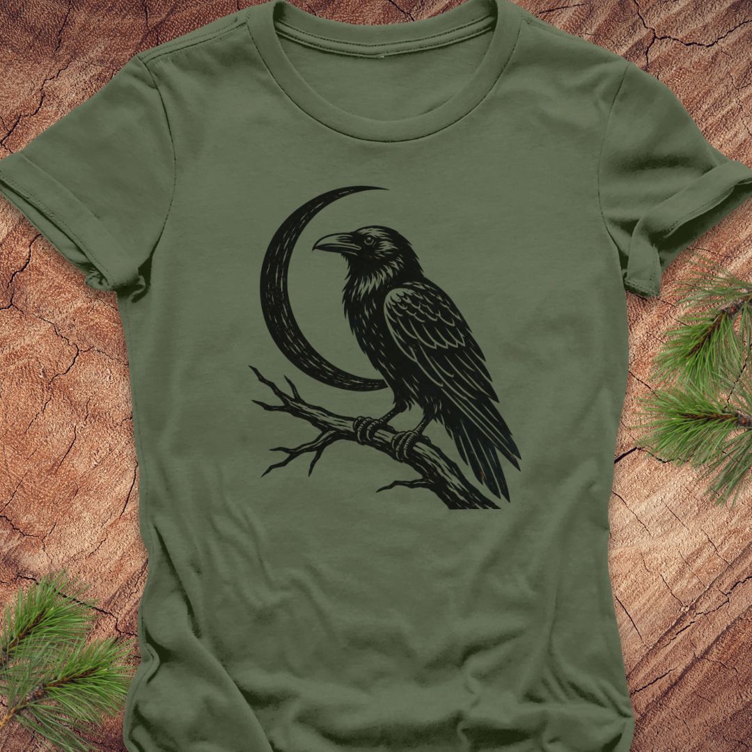 An olive green short-sleeve t-shirt featuring a black graphic of a raven perched on a branch in front of a crescent moon, laid flat on a textured wood background with pine needles in the corners.