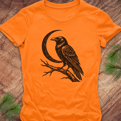An orange short-sleeve crewneck t-shirt featuring a black graphic of a raven perched on a branch next to a crescent moon, laid flat on a rustic wood background with pine needles.