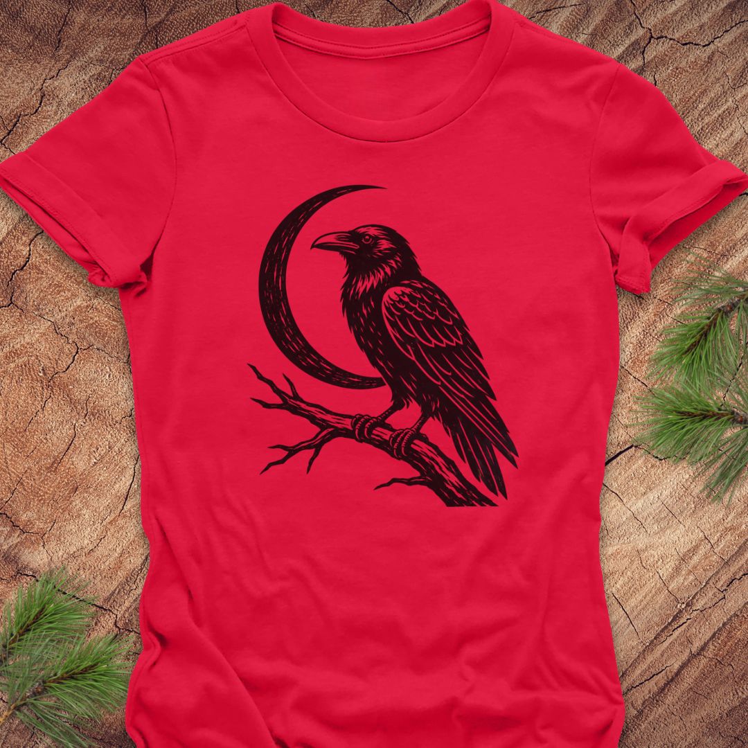 A bright red short-sleeve t-shirt with a black graphic of a raven perched on a branch under a crescent moon, laid flat on a rustic wood surface with pine needles in the corners.