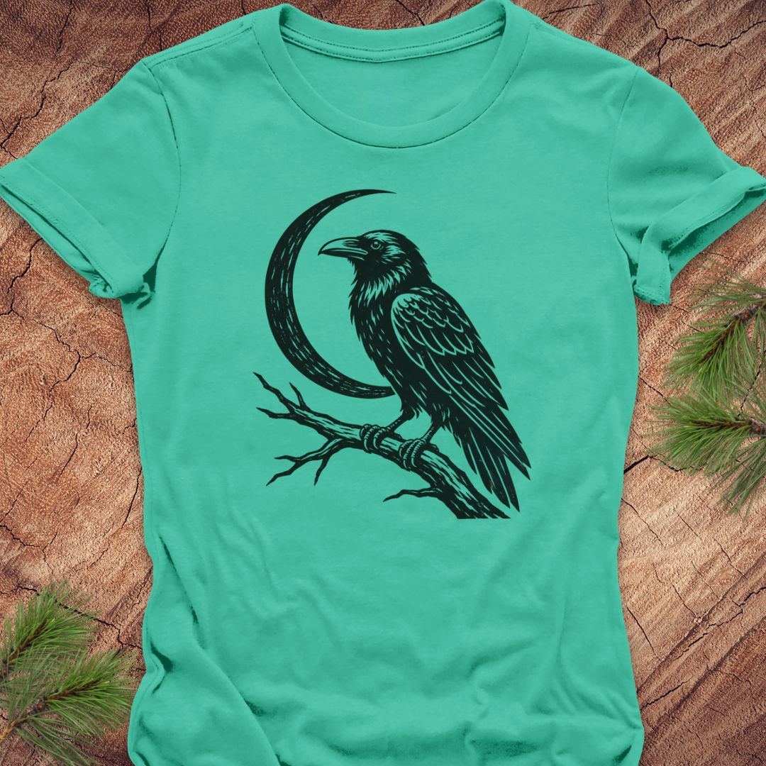A mint green short-sleeve t-shirt with a black graphic of a raven perched on a branch next to a crescent moon, laid flat on a rustic wood background with pine needles.