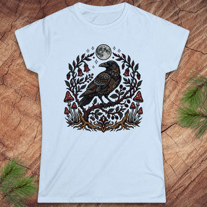 A light blue short-sleeve crewneck t-shirt featuring a detailed graphic of a black raven perched on a branch with a full moon, red mushrooms, and floral elements, laid flat on a rustic wood background with pine needles.