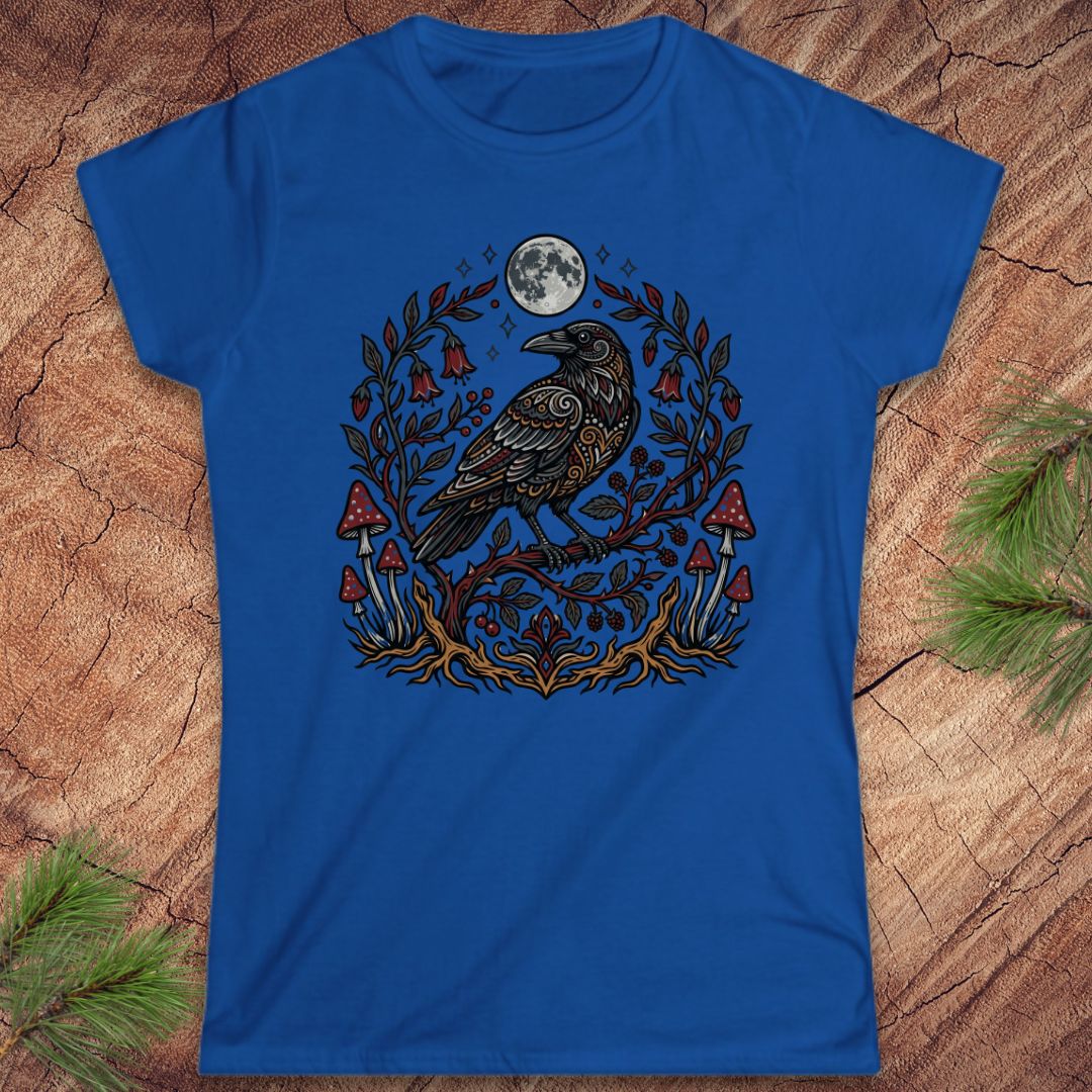 A royal blue short-sleeve t-shirt with a detailed graphic of a raven under a full moon surrounded by mushrooms and foliage, laid flat on a textured wood background with pine branches in the corners.