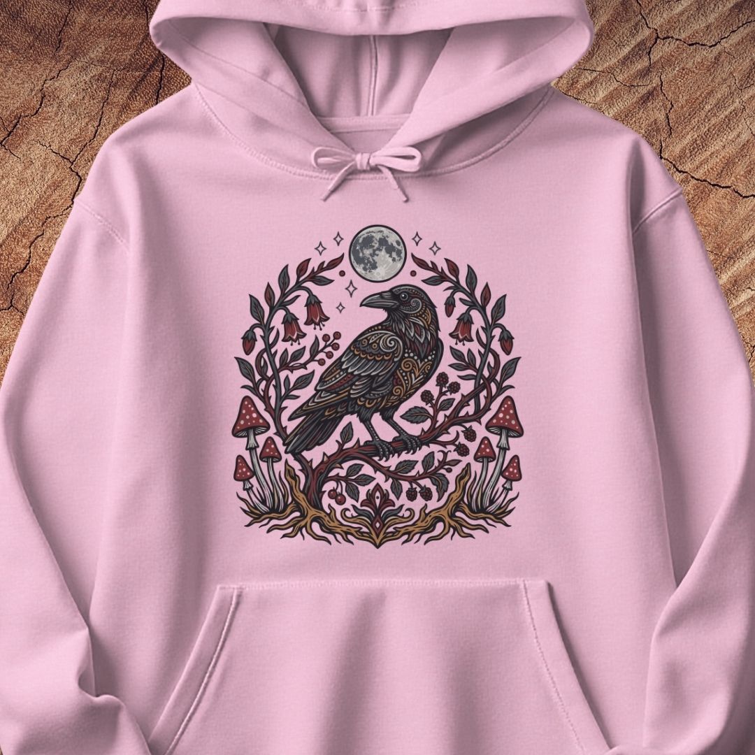 A light pink pullover hoodie featuring a detailed graphic of a raven perched on a branch with a full moon, small bells, and red-and-white mushrooms, displayed on a rustic wood background.