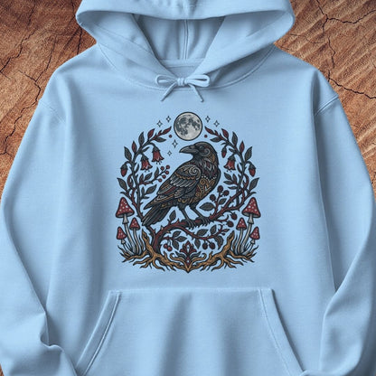 A light blue pullover hoodie laid flat on a textured brown wood background, featuring a detailed graphic of a raven surrounded by a moon, leaves, and red mushrooms on the chest.