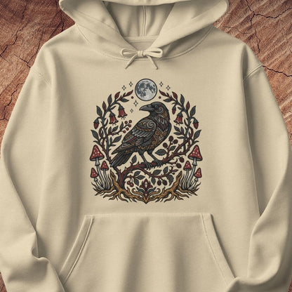 An off-white hooded sweatshirt with a detailed graphic of a black raven perched on a branch surrounded by red mushrooms and green leaves under a full moon, displayed on a textured brown wood background.