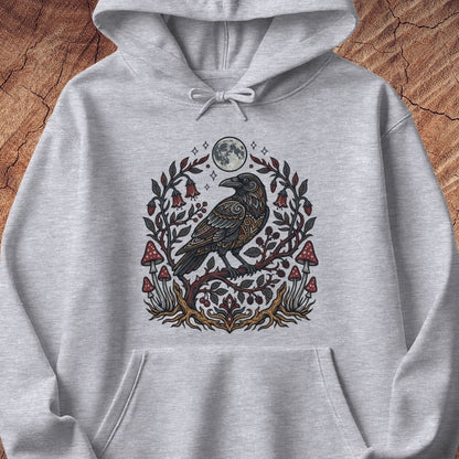 A light heather gray pullover hoodie featuring a detailed graphic print of a raven perched on a branch with red mushrooms, green leaves, and a full moon, displayed flat against a rustic wood background.
