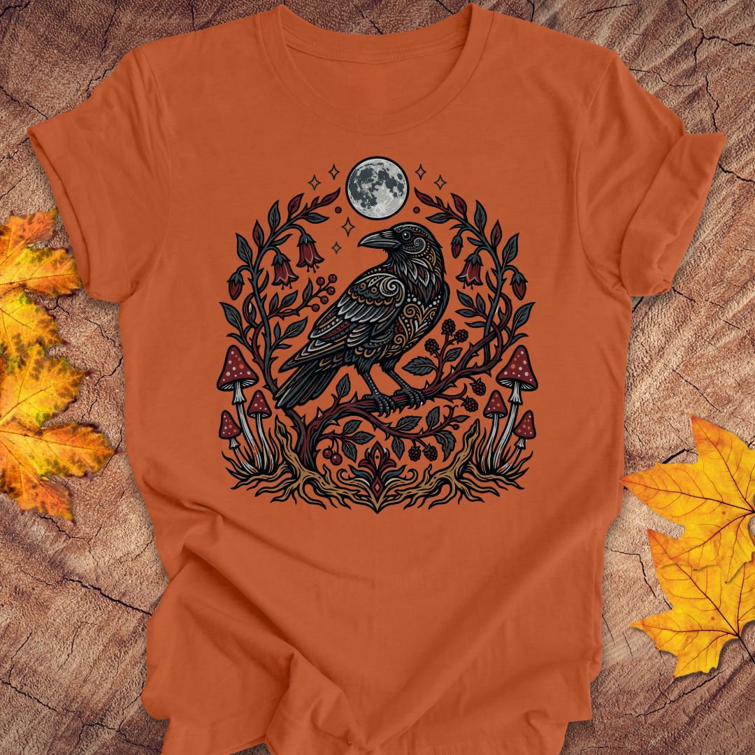 An orange short-sleeve t-shirt with a detailed graphic of a black raven under a full moon, surrounded by a floral wreath and red mushrooms. The shirt is laid flat on a rustic wood background decorated with autumn leaves.