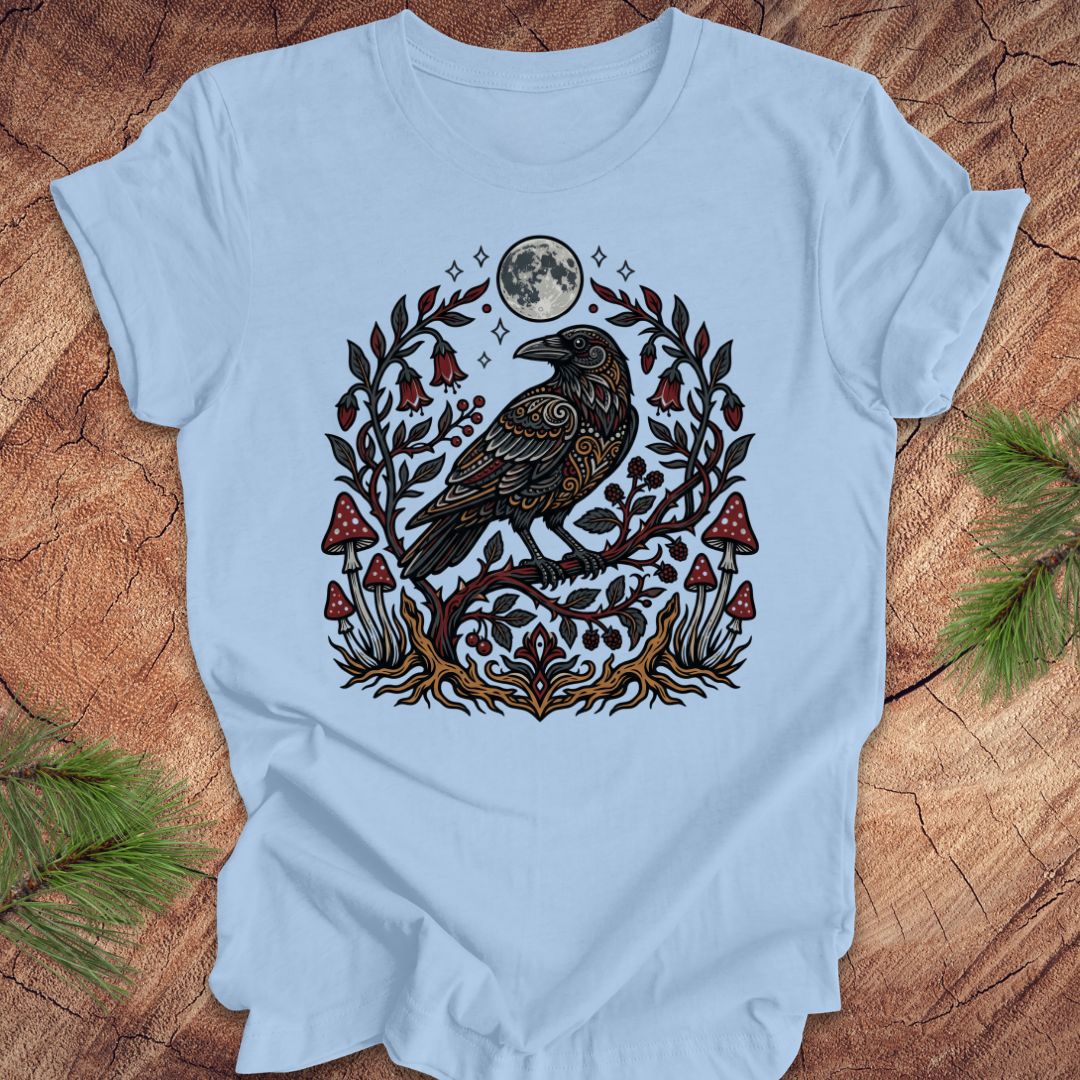 Baby blue t-shirt with a raven and nature-themed design on a wooden surface.