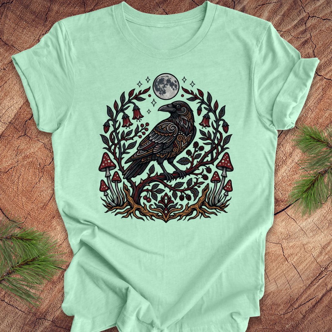 Mint green t-shirt with a raven and nature-themed design on a wooden surface.